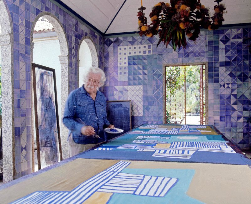 Roberto Burle Marx pintando un mantel en su casa en los años 80. El azulejo de los muros y el candelabro de frutas y flores sobre estructura de y metal son diseños suyos. Fotografía © Tyba 